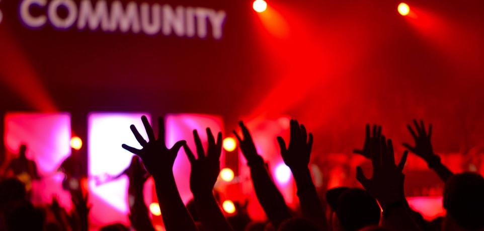 Man sieht die Hände einer feiernden Menschenmenge im rotem Licht. Das Wort 'Community' wird verschwommen an die Wand darüber projiziert.